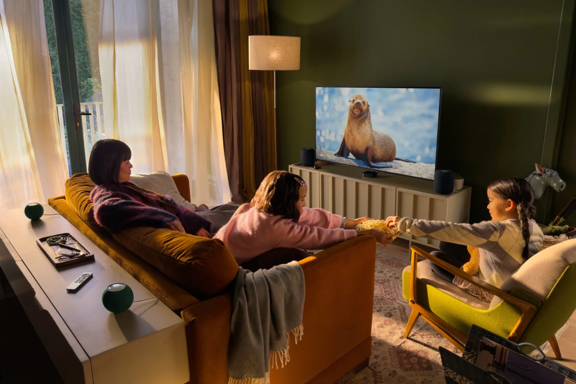 A family watching TV in a living room, with two HomePod speakers beside the TV and two HomePod mini 2 speakers on a table behind the sofa
