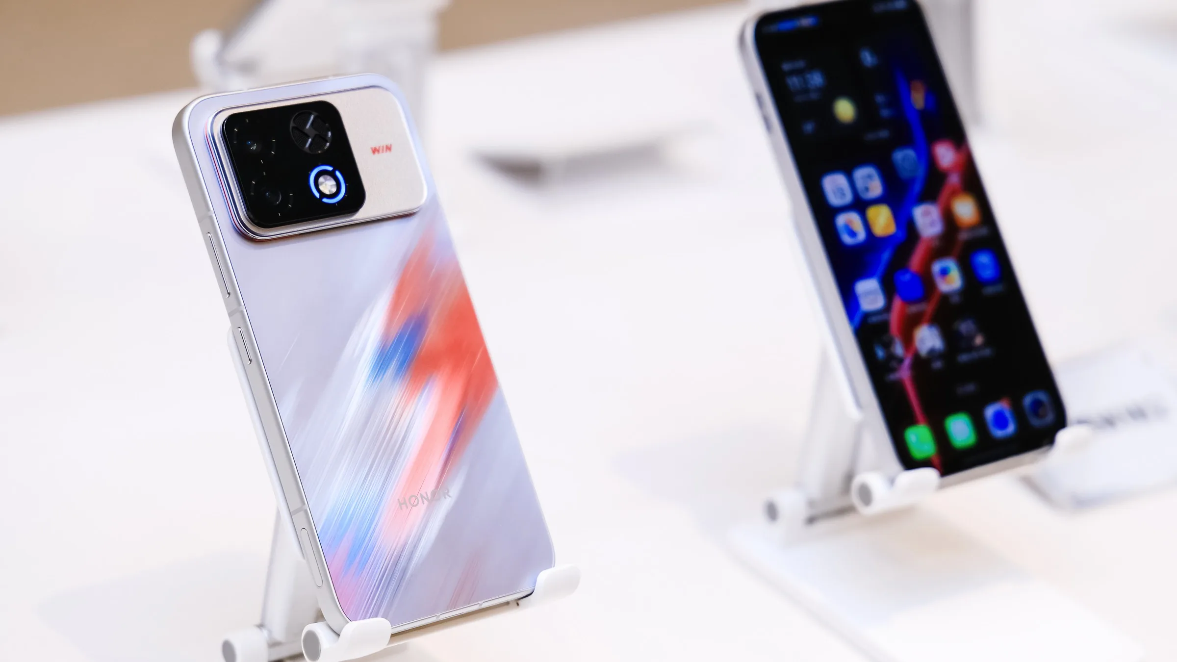 A white Honor Win2 with a red-and-blue streaked back on a display stand, its camera block showing an X-shaped vent and a glowing blue ring, with another phone's screen in the background