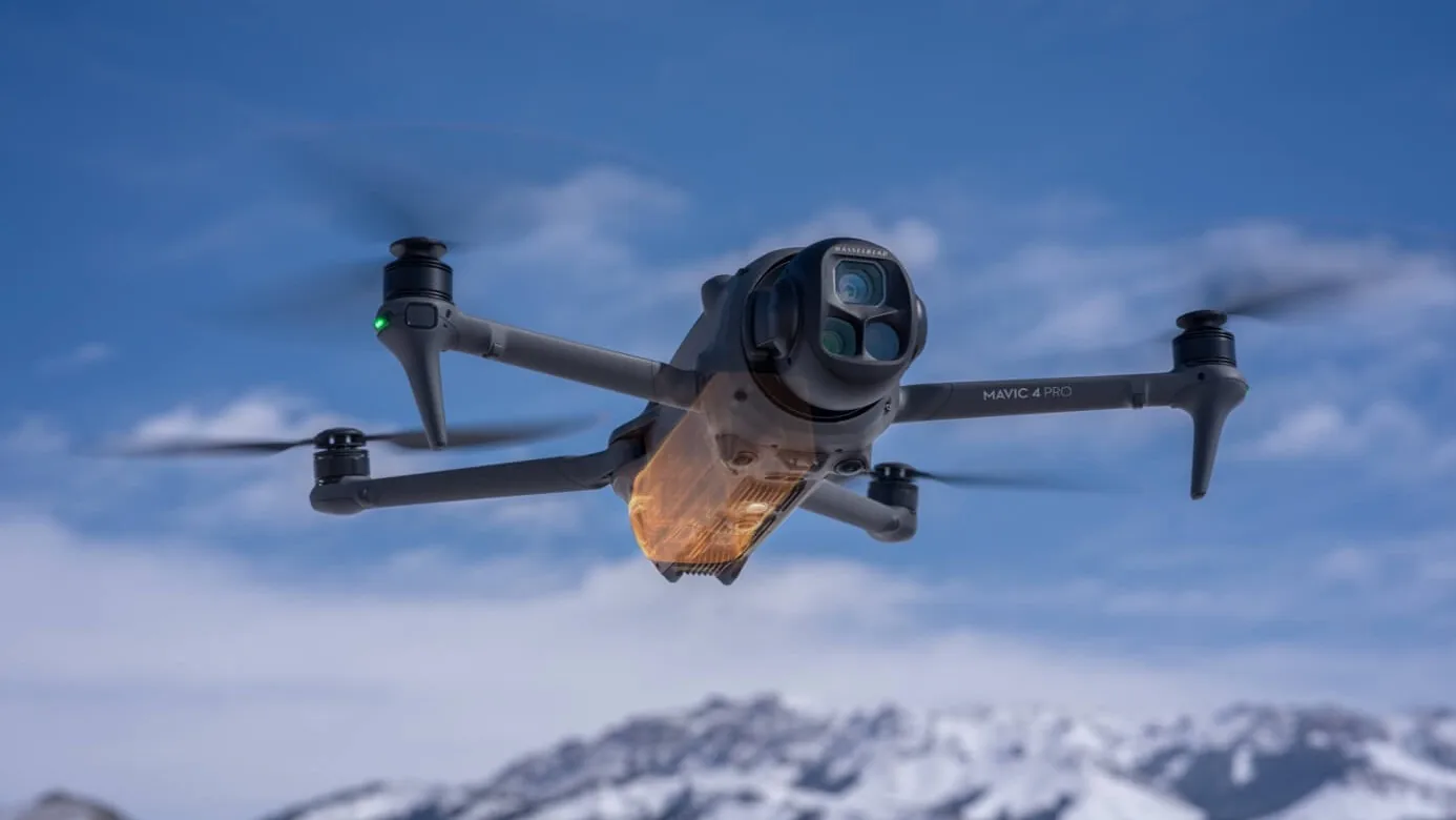 A gray DJI Mavic 4 Pro drone with a large round camera on its nose flies against a blue sky above snowy mountains