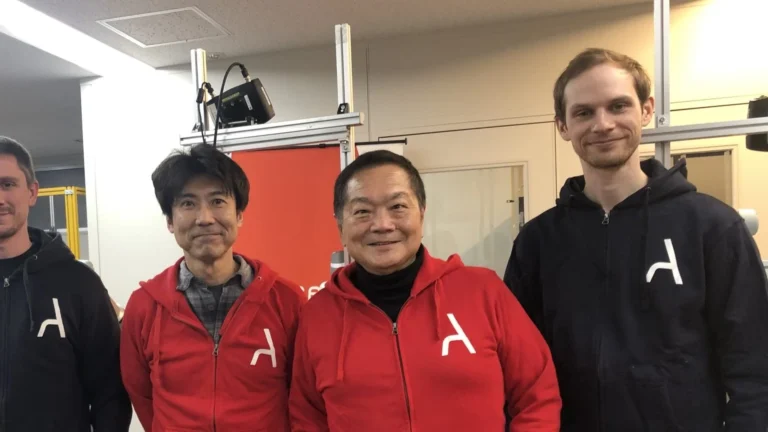 Ken Kutaragi, in a red Ascent hoodie over a black turtleneck, stands smiling with three other people in Ascent hoodies in a robotics lab