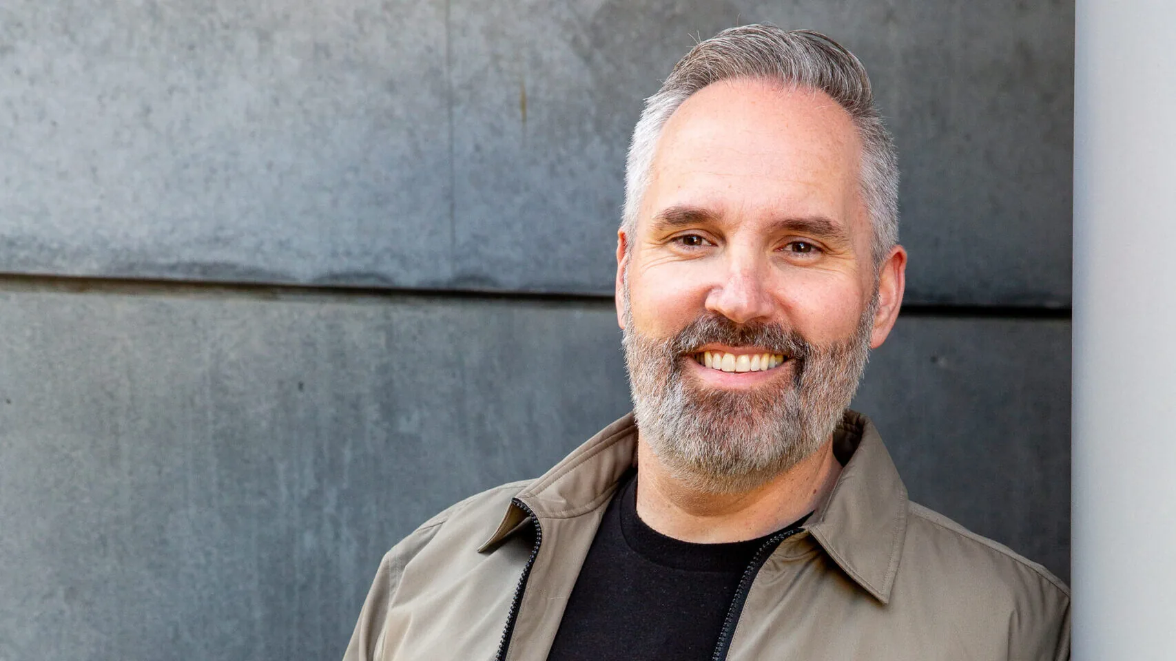 Scott Van Vliet, smiling, in an olive jacket over a black T-shirt, against a gray stone wall