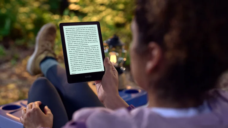 A person with their feet up outdoors reading a Kindle Paperwhite, with a lantern glowing in the background