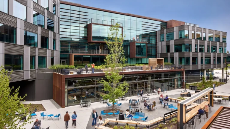 Glass and steel office buildings around a courtyard with blue chairs, tables, wooden benches and people talking, walking and working