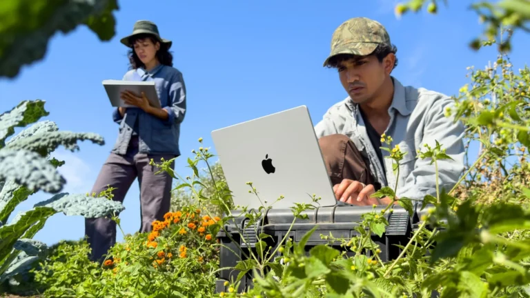 A man works on a silver 14-inch MacBook Pro set on a case in a garden, while a colleague takes notes behind him
