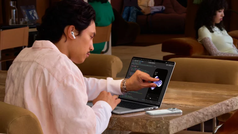 A man seated at a table touches the screen of the new MacBook Pro with PROLED XDR Display while it shows an app beside code