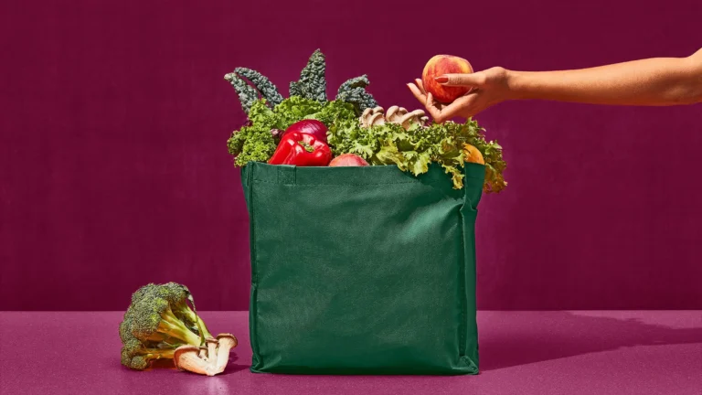 A green reusable bag full of kale, lettuce and a red pepper, with a hand adding a peach, on a maroon background