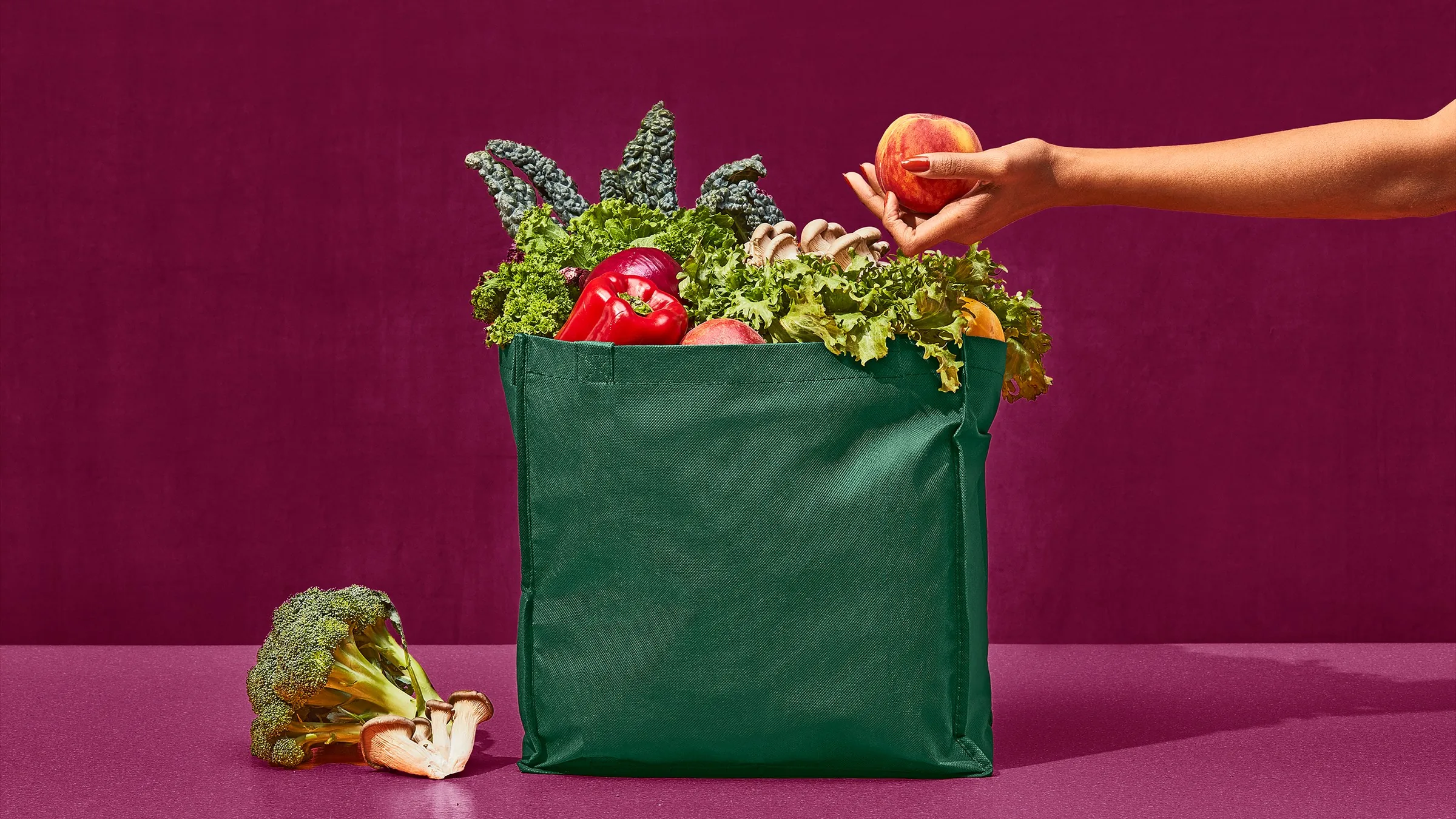 A green reusable bag full of kale, lettuce and a red pepper, with a hand adding a peach, on a maroon background