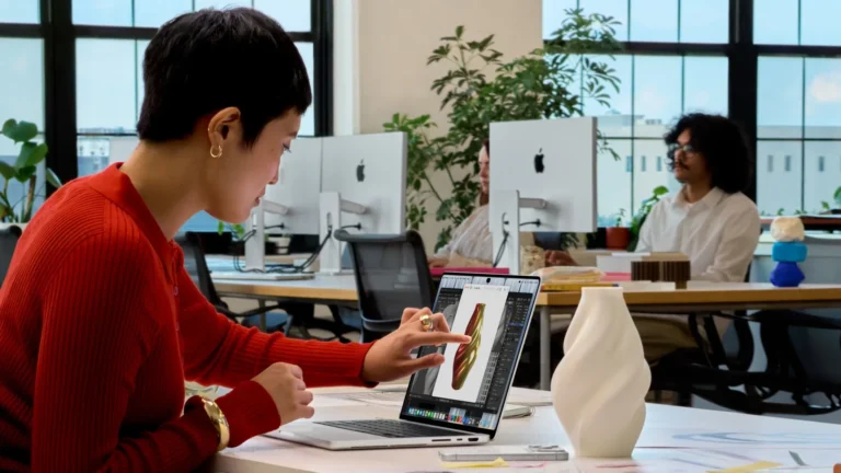 A woman in a red sweater touches a 3D model of a vase on the screen of a silver MacBook Pro with PROLED XDR Display at an office desk, with a 3D-printed white vase beside it