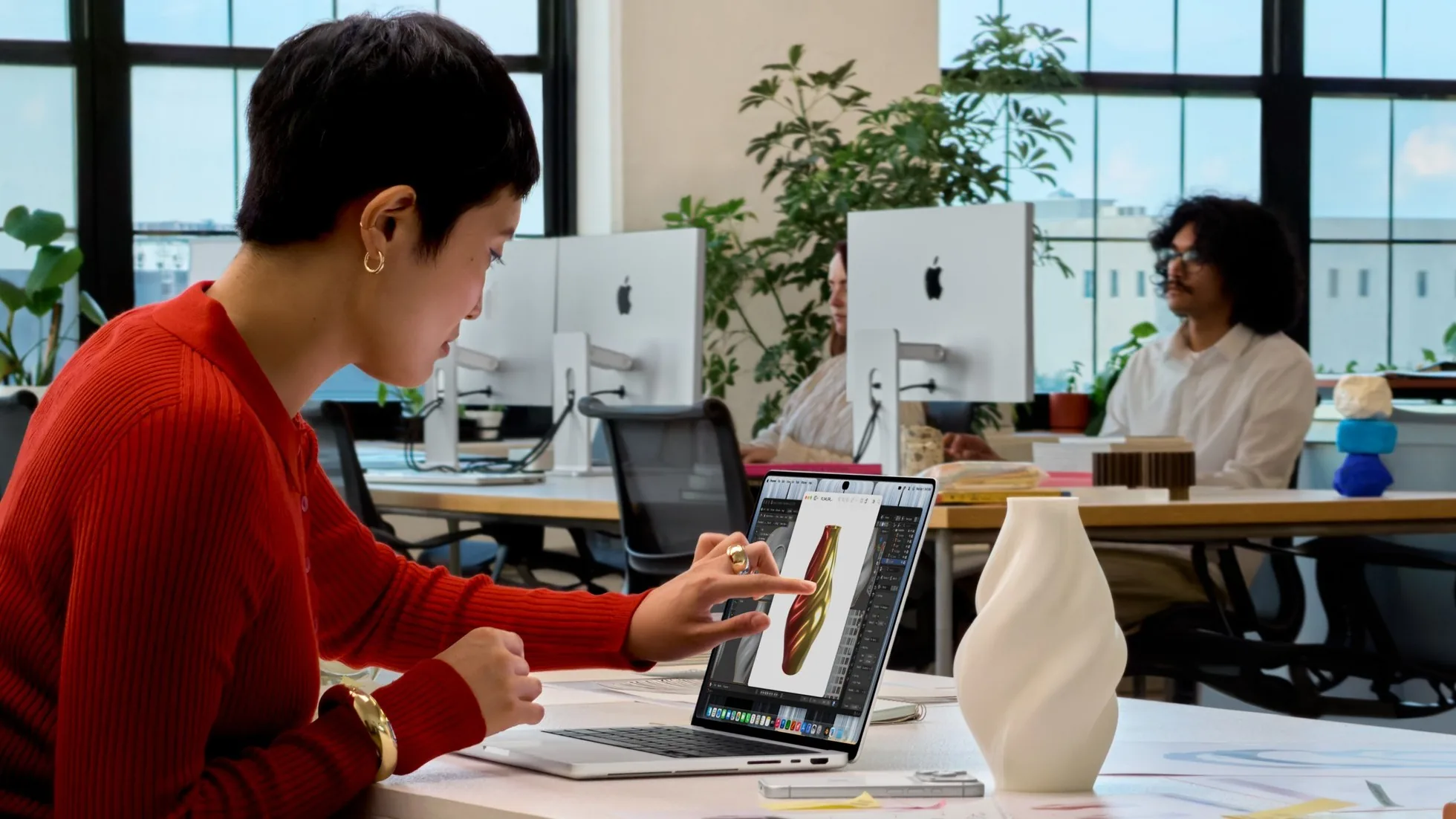 A woman in a red sweater touches a 3D model of a vase on the screen of a silver MacBook Pro with PROLED XDR Display at an office desk, with a 3D-printed white vase beside it