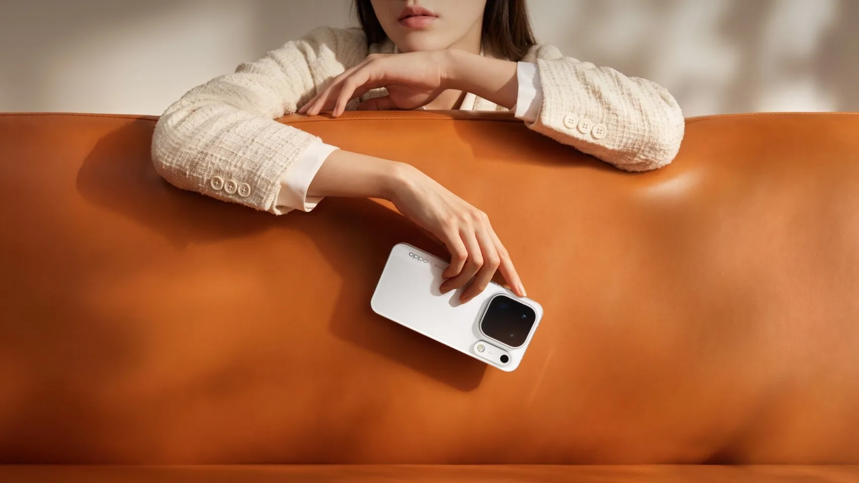 A woman leans on the back of an orange leather sofa, holding a white Oppo Find X10 Pro Max with its camera module facing up