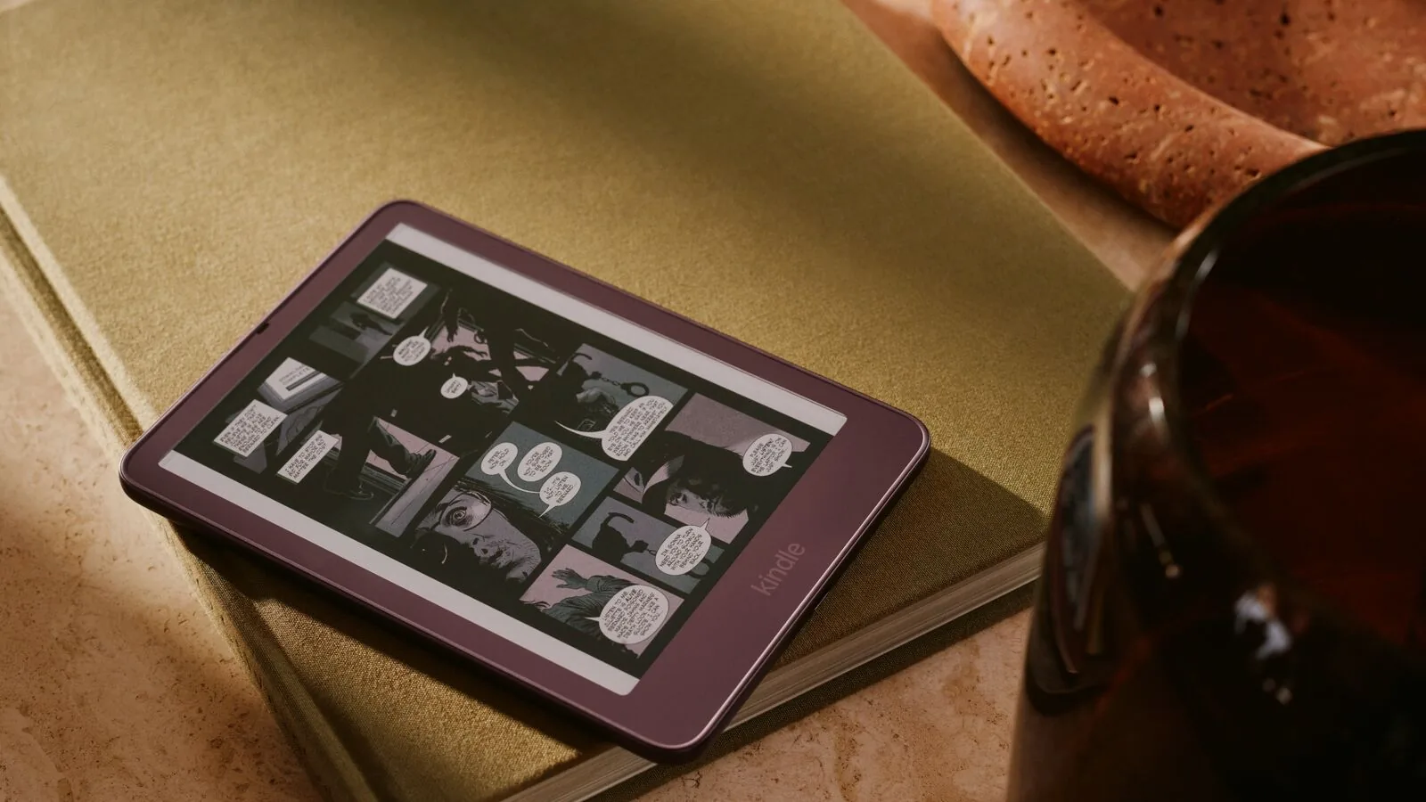 A purple-red Kindle Colorsoft lying on a green cloth-bound book, its screen showing a comic page in muted color