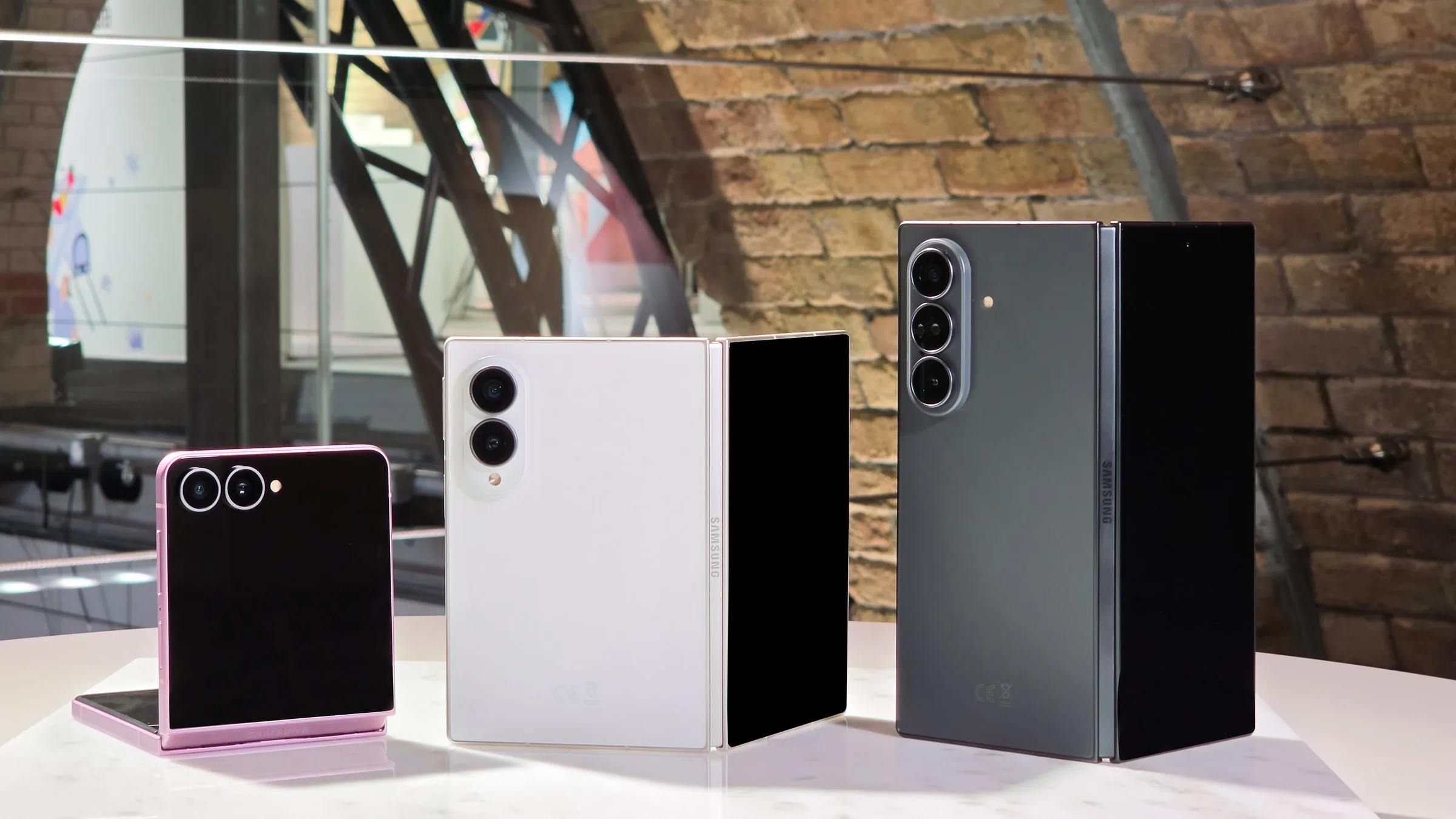 Three Samsung foldable phones standing on a white table in front of a brick wall: a pink Galaxy Z Flip8, a white Galaxy Z Fold8 and a gray Galaxy Z Fold8 Ultra, each half open.
