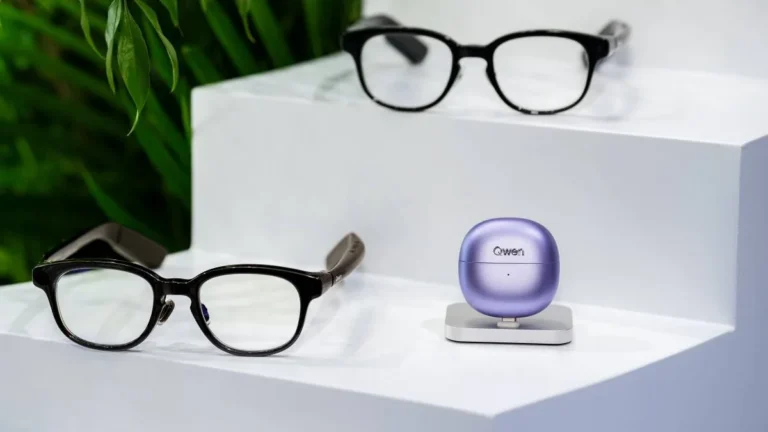 Two pairs of black-framed Qwen Glasses on white display steps beside a lilac Qwen Clip earbuds case on a silver stand