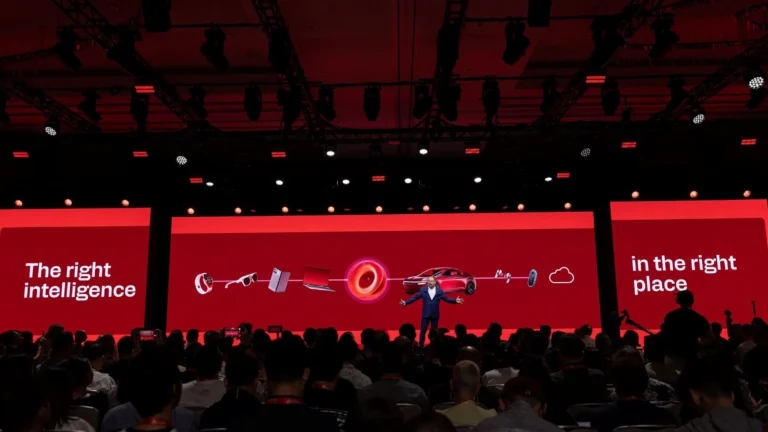 Cristiano Amon on stage with his arms spread in front of a red screen showing a watch, glasses, a phone, a laptop, a car and earbuds, with a crowd in the foreground