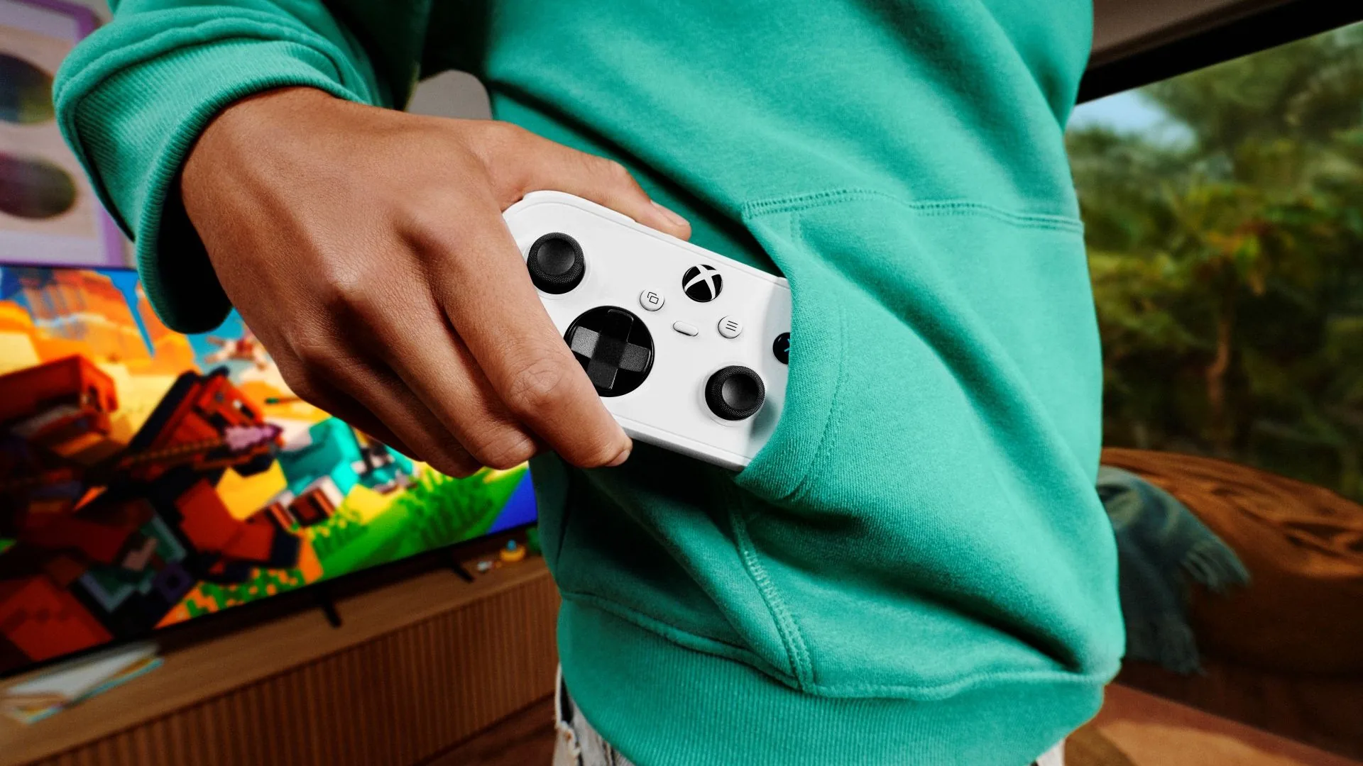 A hand slipping a small white Xbox Nano Controller into the front pocket of a green hoodie, with a TV in the background