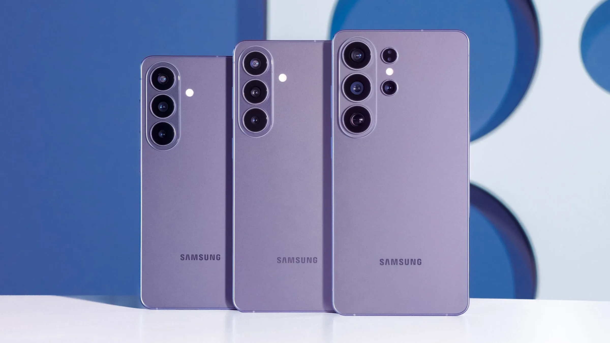 Three Samsung phones in a purple shade seen from the back, standing upright on a white table in front of a blue and white backdrop: from left, Galaxy S26, Galaxy S26+ and Galaxy S26 Ultra.