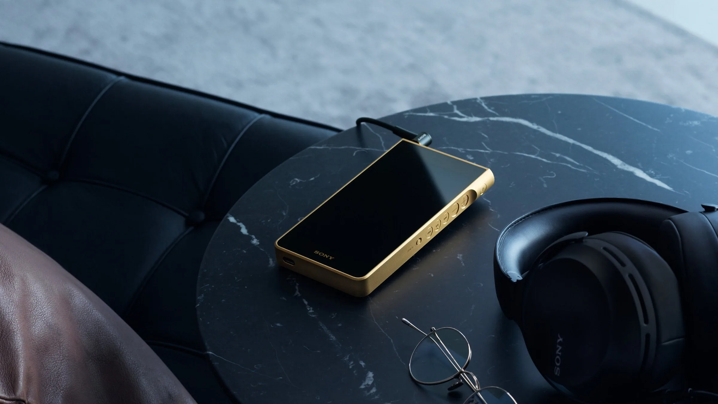A gold Sony NW-WM1ZM3 Walkman on a black marble table beside a pair of Sony headphones and reading glasses